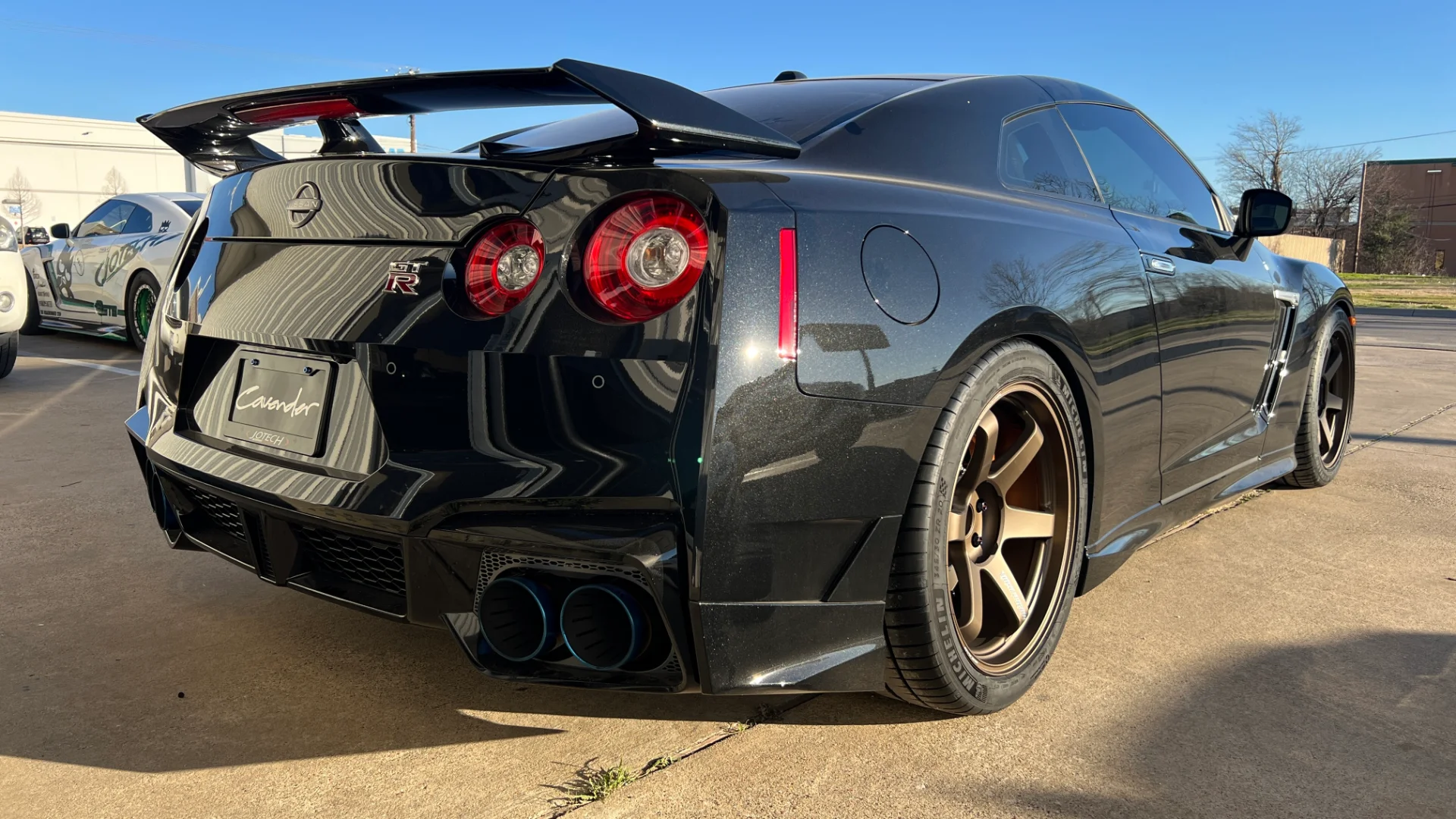 Rear view of a black Nissan GT-R with aftermarket bronze wheels, a large rear wing, and blue exhaust tips.