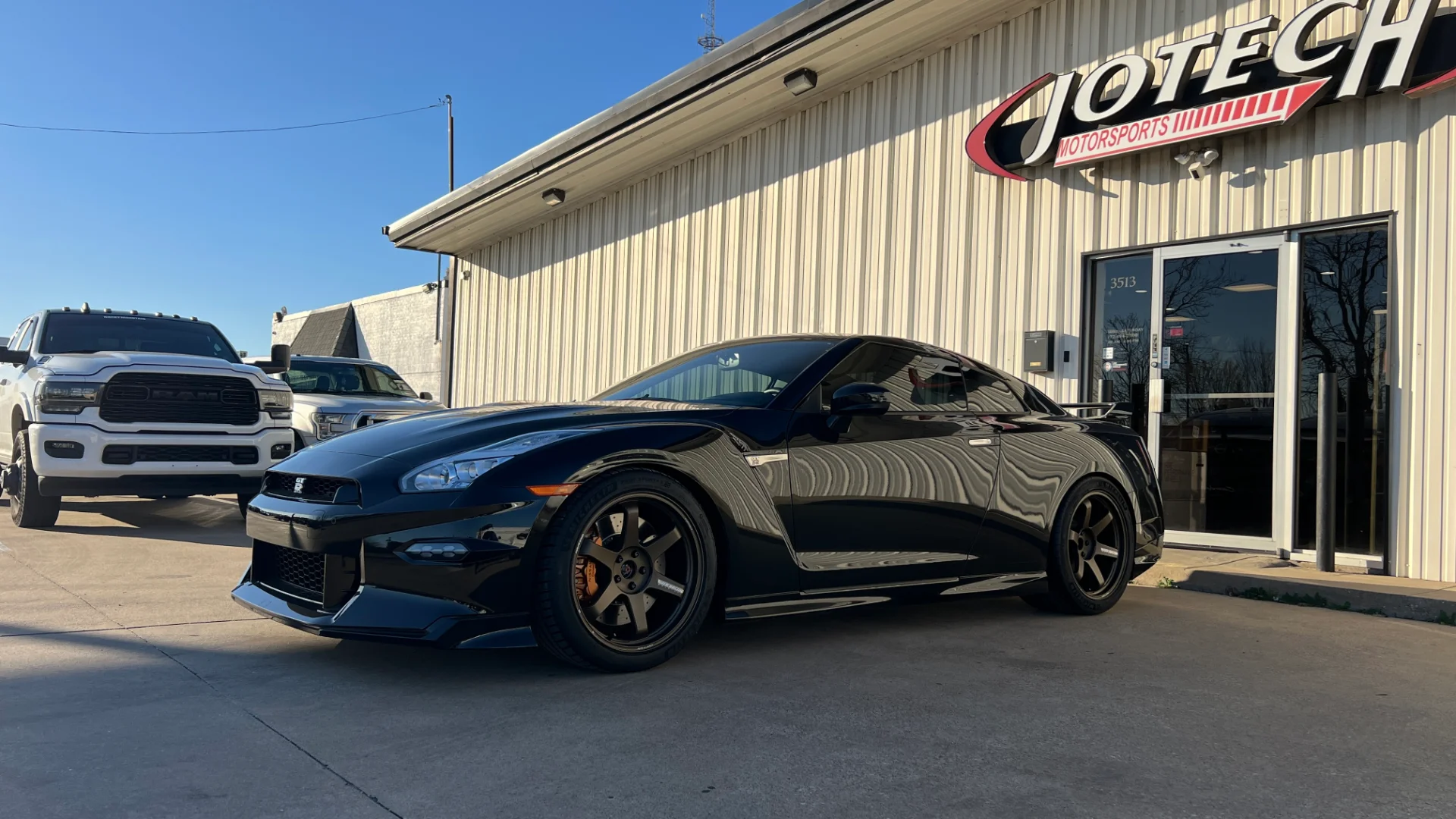 A black Nissan GT-R parked outside the Jotech Motorsports performance tuning shop with its prominent signage visible.