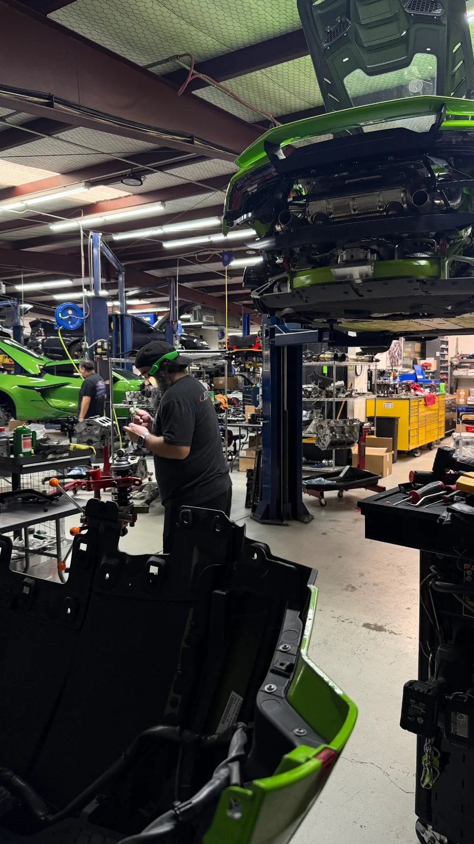 A green Lamborghini on a lift in a busy auto repair shop, with mechanics working on vehicles and parts.