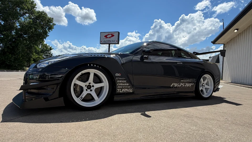 A black Nissan GT-R with a widebody kit, aftermarket wheels, and a large rear wing parked outside a Jotech Motorsports facility.
