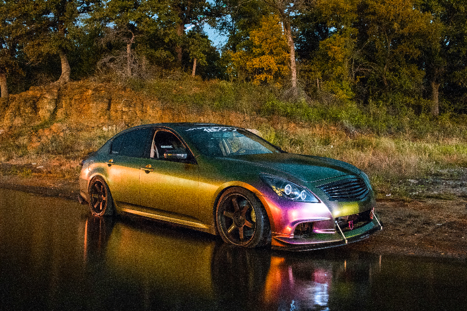 A custom Infiniti G37 sedan with a chameleon wrap, aftermarket wheels, and custom headlights parked on a wet road at dusk.