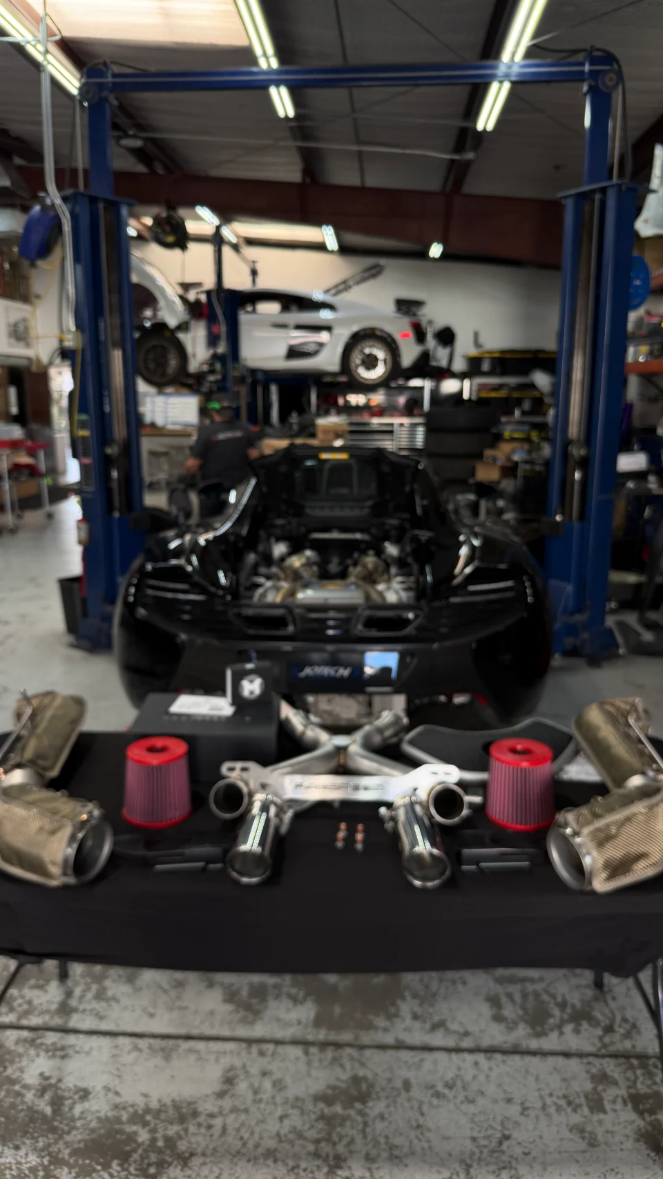 Aftermarket exhaust system and air filters laid out on a table in a Jotech Motorsports shop with a black McLaren and white Audi R8 on lifts in the background.