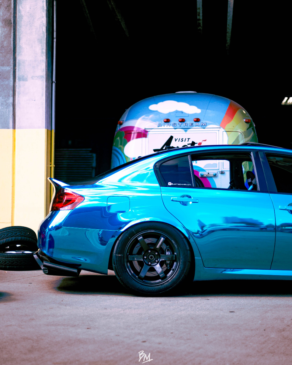 A chrome blue Infiniti G37 sedan with aftermarket wheels and a rear diffuser parked in front of a colorful Airstream trailer.
