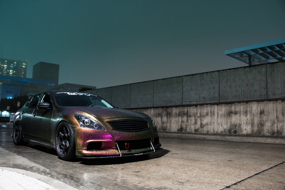 A custom chameleon-painted Infiniti G37 sedan with aftermarket wheels and a front splitter parked in an urban setting at night.