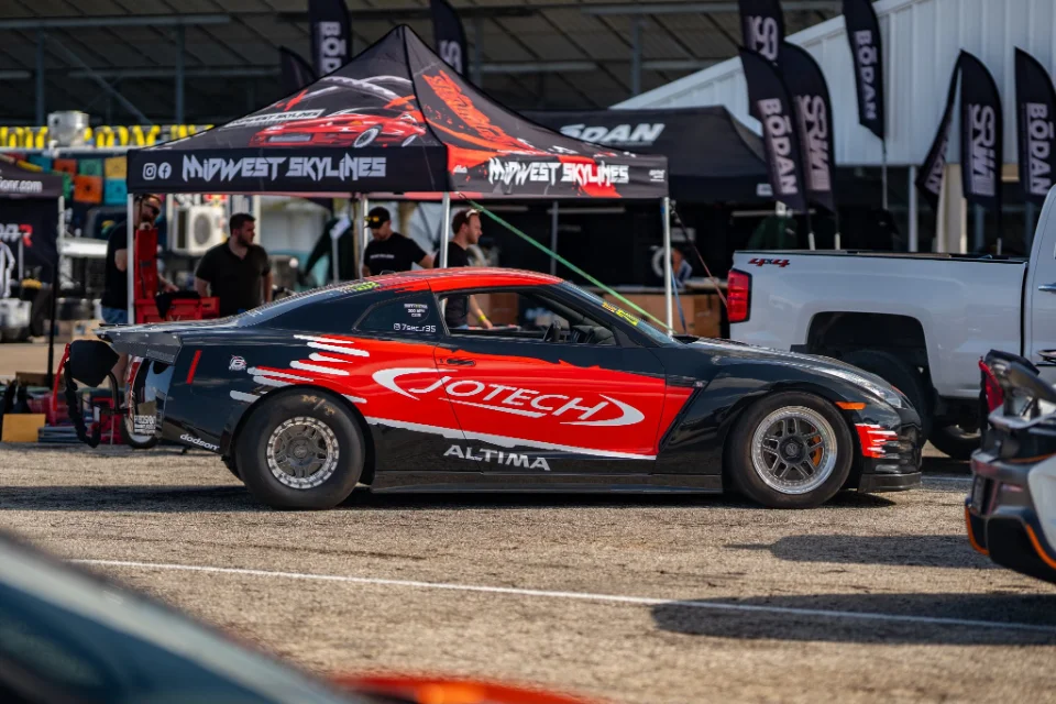 A black and red Jotech Nissan GT-R drag car with widebody and drag slicks parked at a track event.