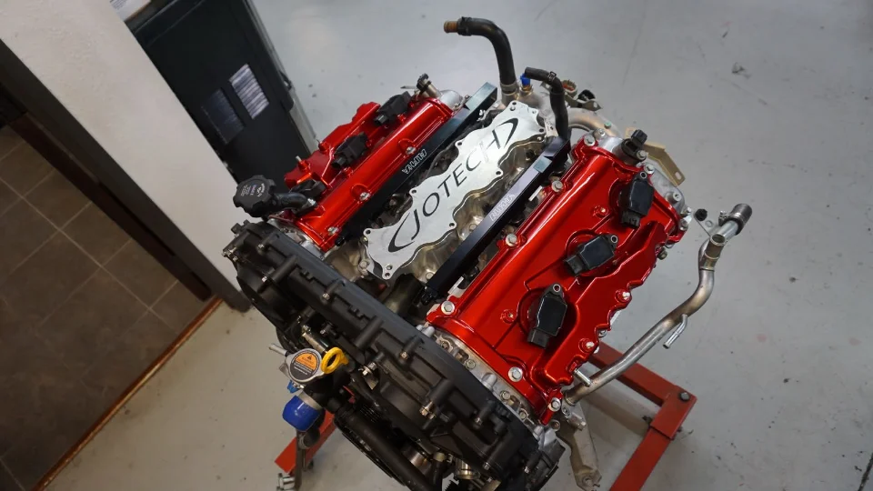 A detailed view of a Jotech built engine with custom red valve covers and a polished intake manifold cover on an engine stand.