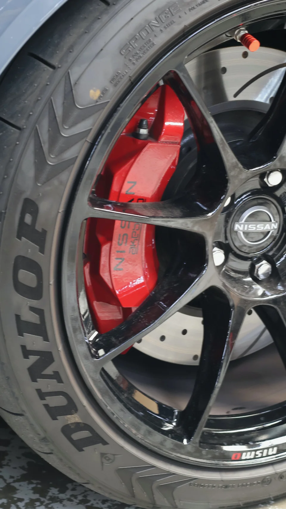A close-up of a Nissan car's black multi-spoke wheel with red Nismo brake calipers and Dunlop tires.