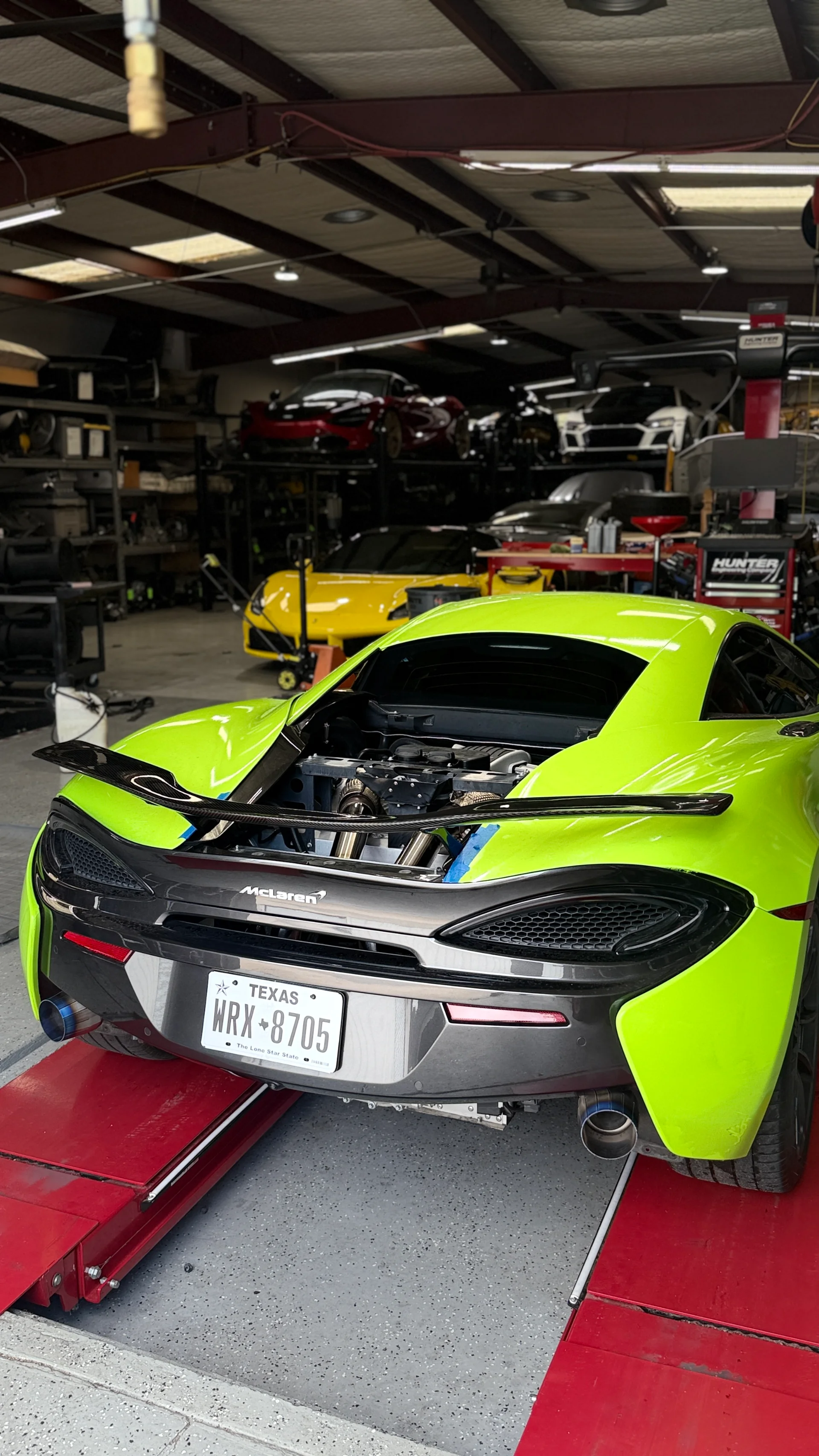 A lime green McLaren 570S with its engine bay open, revealing a custom turbo and exhaust system, inside a performance tuning shop.