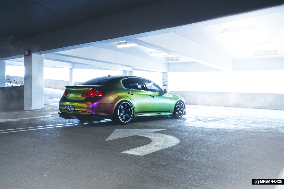 A custom Infiniti G37 sedan with a vibrant chameleon color-shift wrap and aftermarket wheels parked in a bright concrete parking garage.