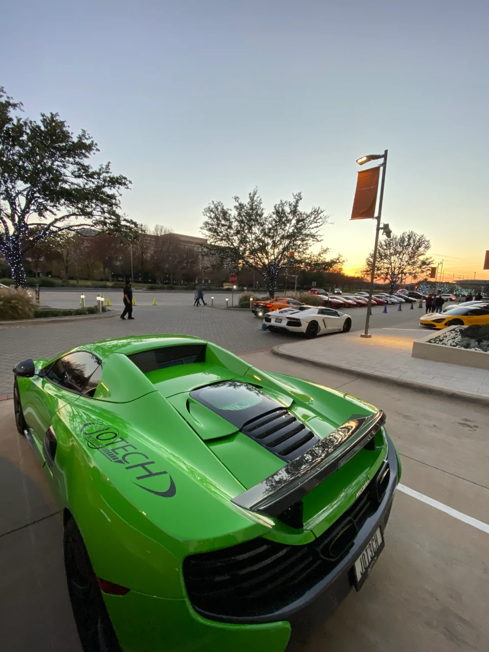A vibrant green McLaren with Jotech branding parked at an outdoor supercar meet during sunset, with other luxury cars in the background.