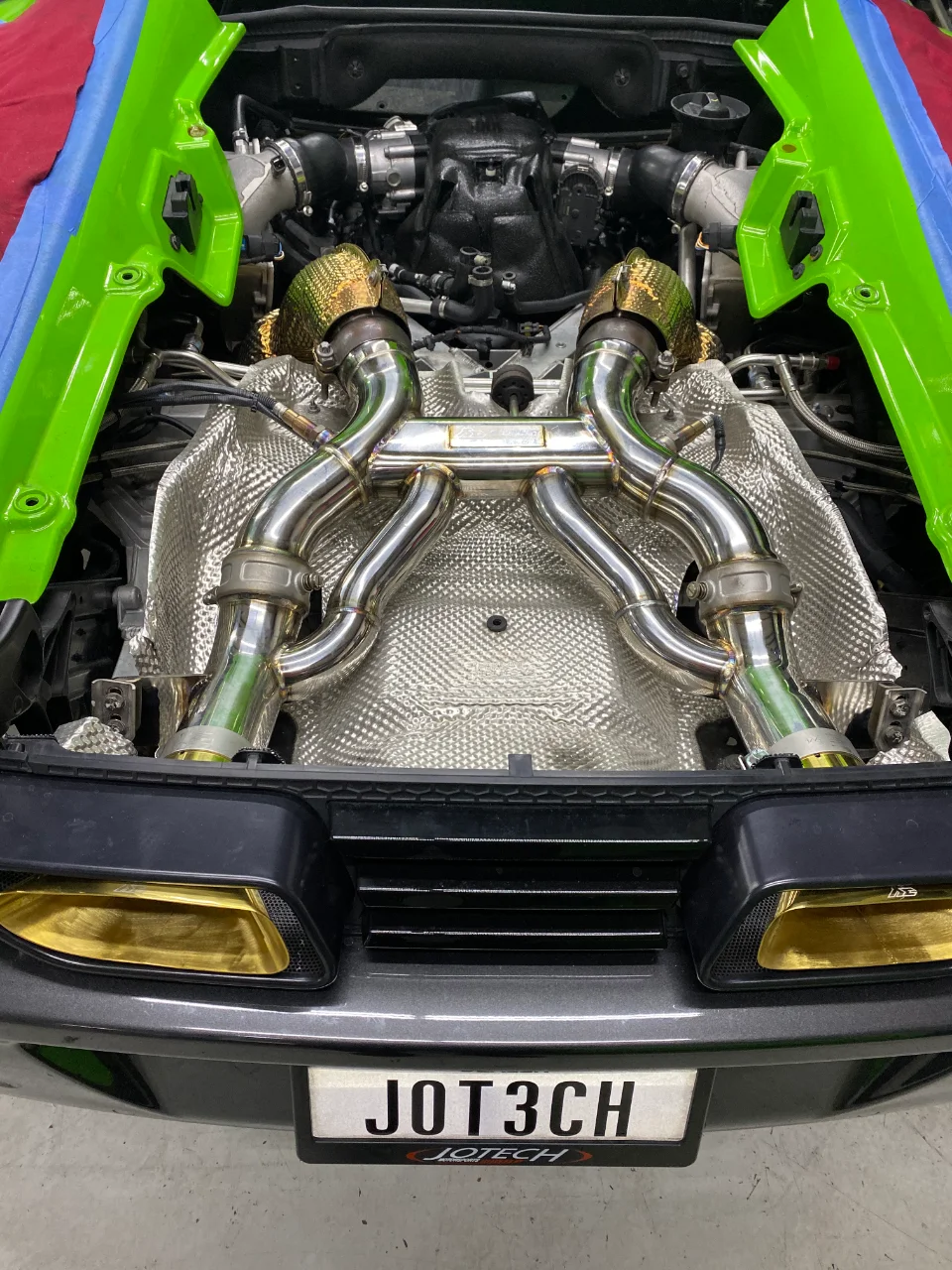 A close-up view of a Lamborghini Huracan engine bay showcasing a polished aftermarket exhaust system and turbochargers.