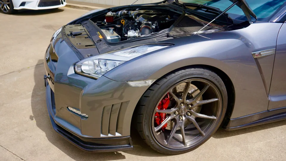 A grey Nissan GT-R with its hood open, showcasing the engine bay with aftermarket induction, custom wheels, and red brake calipers.