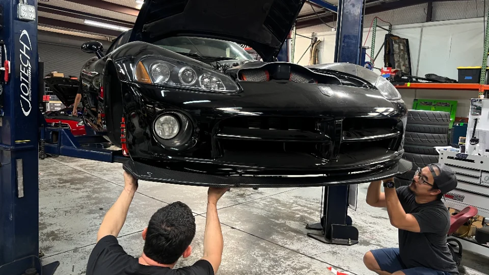 Two mechanics install a carbon fiber splitter on a black Dodge Viper on a lift in the Jotech Motorsports shop.