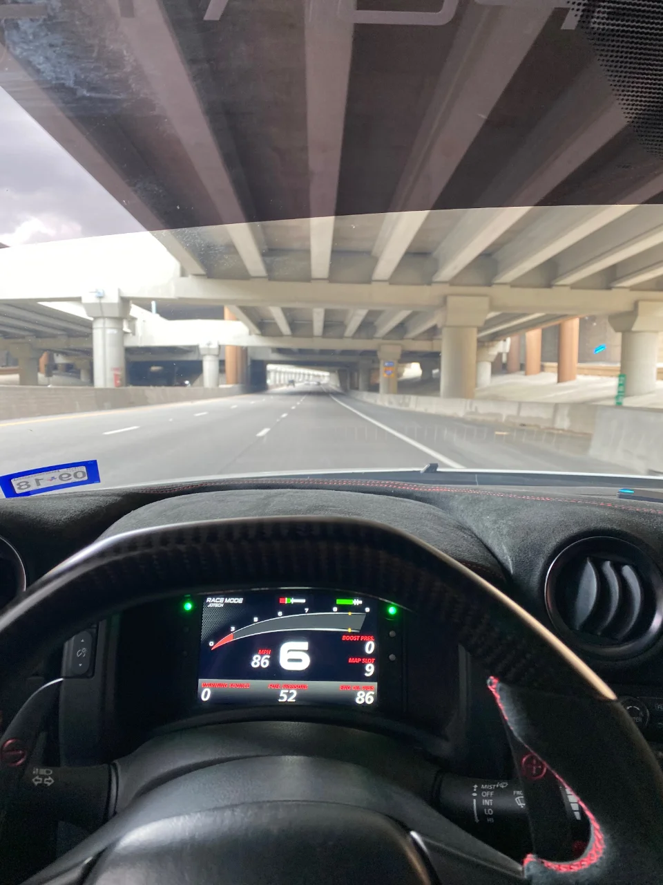 Driver's perspective from a performance car interior, showing a custom steering wheel and digital dashboard displaying 86 MPH while driving on a highway.