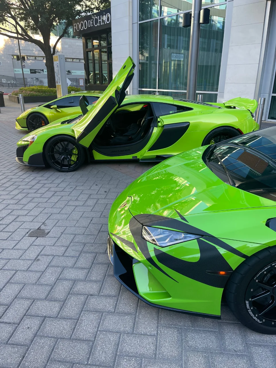Three lime green supercars, including a McLaren and a Lamborghini, parked outside a building with Fogo de Chao signage.