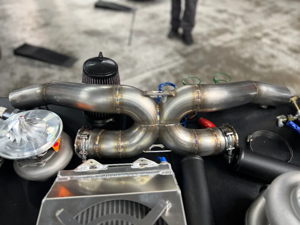 A display of high-performance car parts featuring a stainless steel exhaust X-pipe, turbocharger, and air filter.