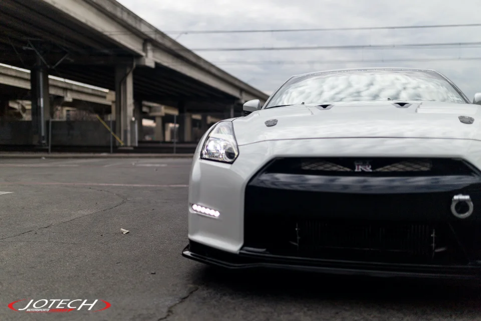 White Nissan GT-R R35 with aftermarket front splitter and LED daytime running lights parked under an overpass.