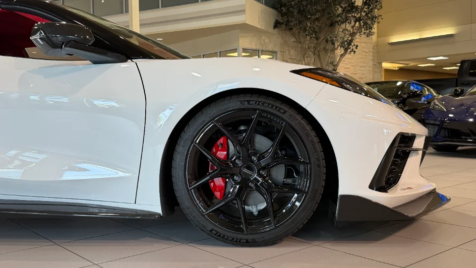 Close-up of a white C8 Corvette's front wheel with black aftermarket wheels and red brake calipers in a showroom.