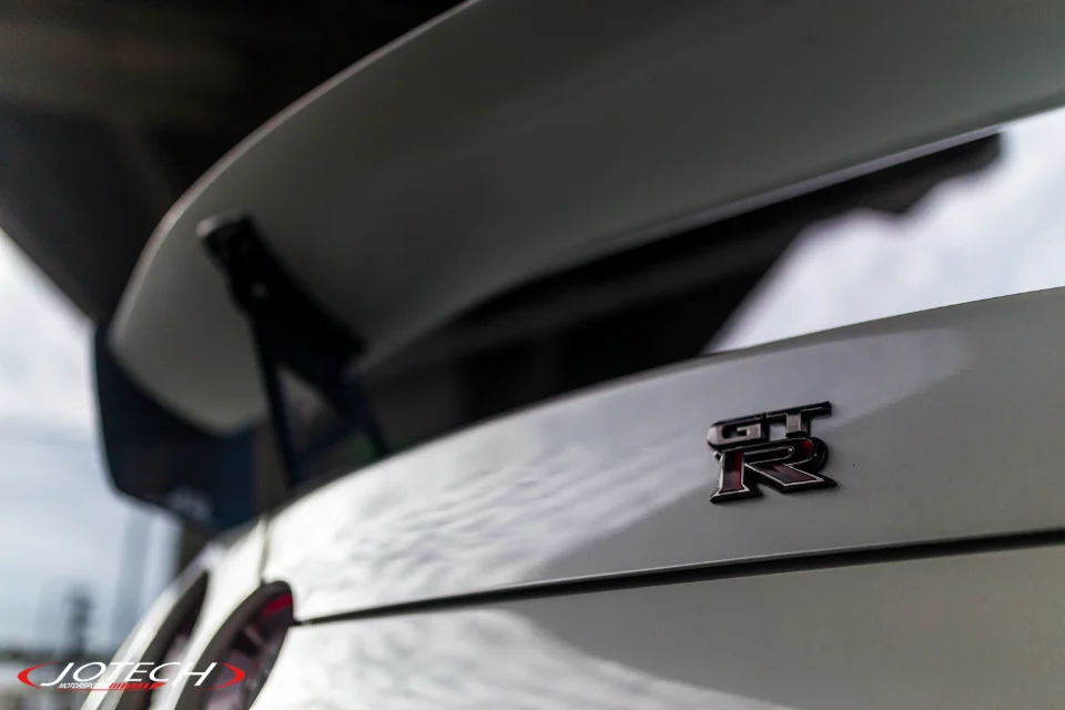 A close-up of the rear of a white Nissan GT-R, highlighting the GT-R emblem and large rear wing.