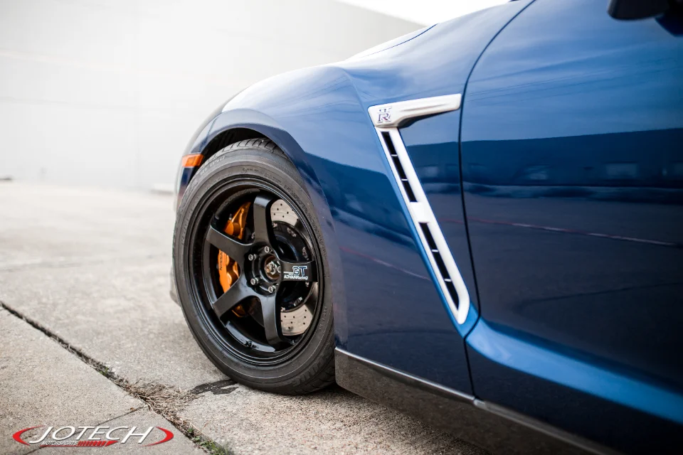 A close-up detail shot of a blue Nissan GT-R's front wheel, black aftermarket wheels, and orange brake calipers.
