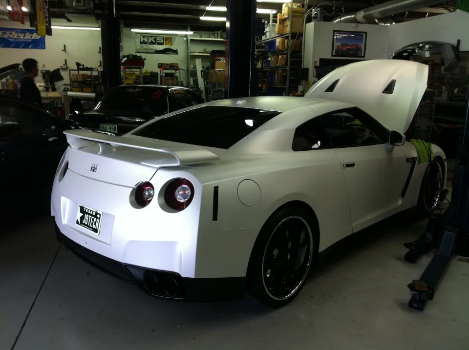 A white Nissan GT-R with its hood open in a Jotech Motorsports shop, with a car lift nearby.