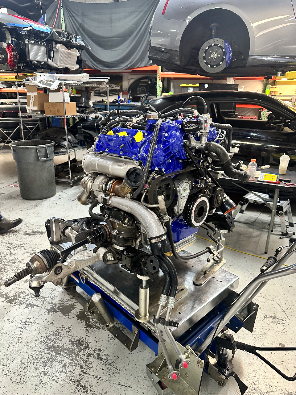A high-performance twin-turbo engine assembly with blue valve covers and intake manifold on a stand in a Jotech Motorsports shop.