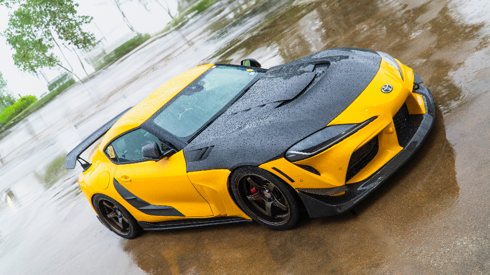 A modified yellow and black Toyota Supra with a carbon fiber hood, large rear wing, and aftermarket wheels parked on a wet surface.