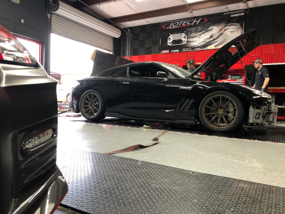 A black Nissan GT-R with its front bumper removed, exposing an intercooler, being worked on by mechanics in a tuning shop.