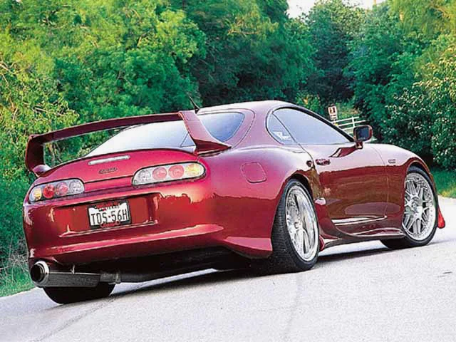 Red Toyota Supra MK4 with aftermarket wheels and a large rear wing parked on a road with trees in the background.