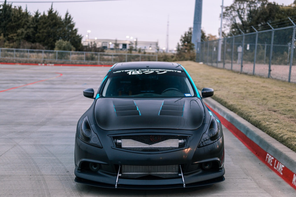 Front view of a matte black Infiniti G37 with a vented hood, large intercooler, and front splitter, parked outdoors.