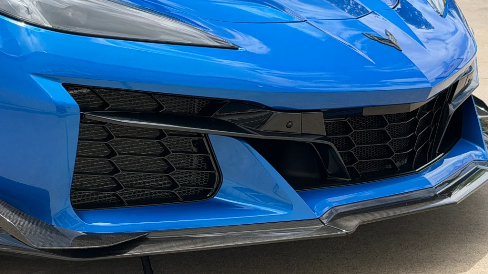 A close-up of a blue C8 Corvette's front bumper, featuring black honeycomb grilles and a carbon fiber splitter.