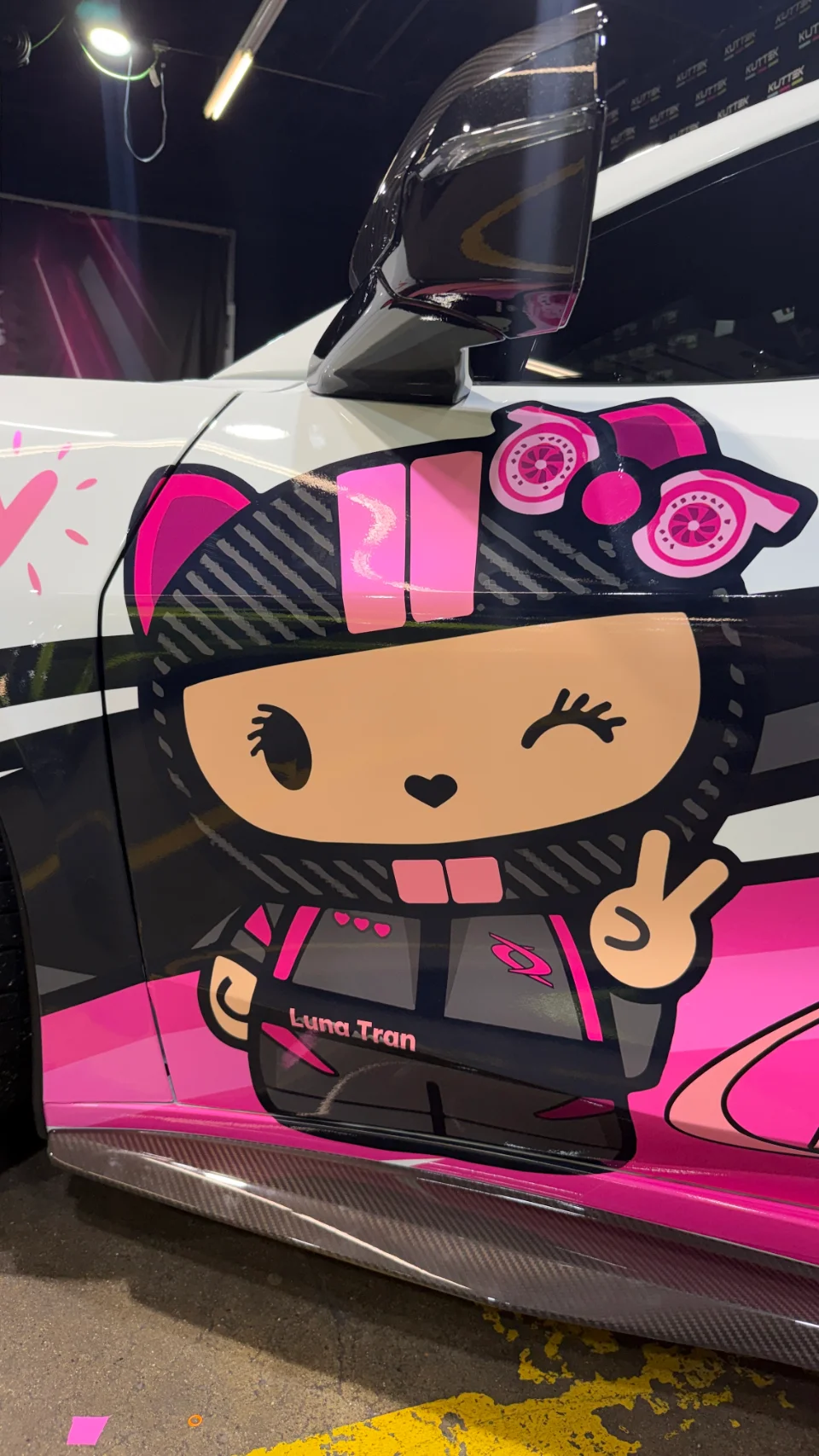 Close-up of a car's side with a vibrant pink and black custom vinyl wrap featuring a cartoon character and a carbon fiber side skirt.