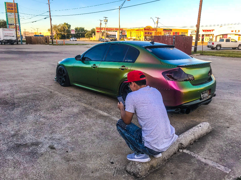 An Infiniti G37 sedan with a chameleon wrap and aftermarket wheels parked in a lot, with a person crouching to take a photo.