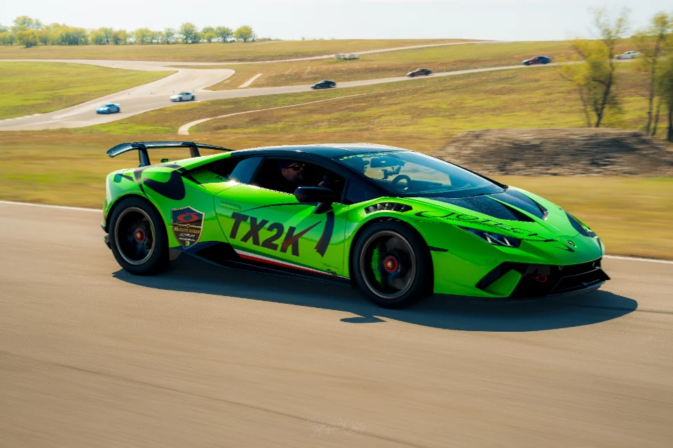 A vibrant green Lamborghini Huracan with TX2K and Jotech livery speeds around a race track under a clear sky.