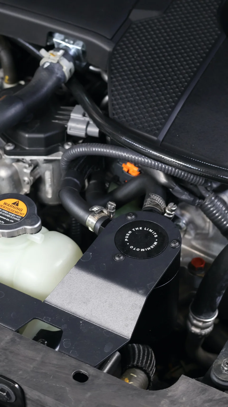 Close-up of a Mishimoto oil catch can installed in a car's engine bay, showing hoses and other engine components.