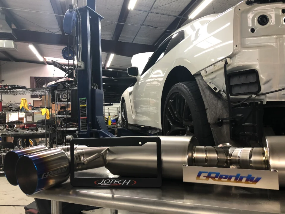 A GReddy exhaust system in the foreground with a white Nissan GT-R on a lift in the Jotech Motorsports shop.