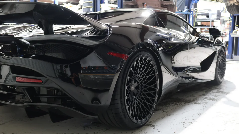 A sleek black McLaren 720S with custom black multi-spoke wheels and a prominent rear wing, parked inside a shop.