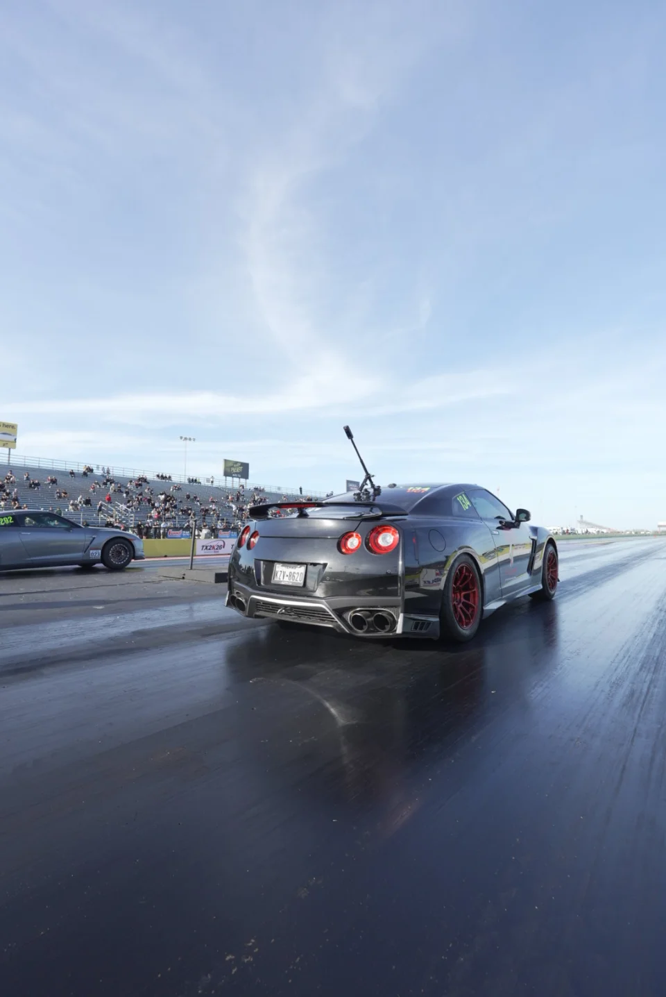 Rear view of a black Nissan GT-R with red wheels at the drag strip starting line, with another car in the left lane and spectators in the stands.