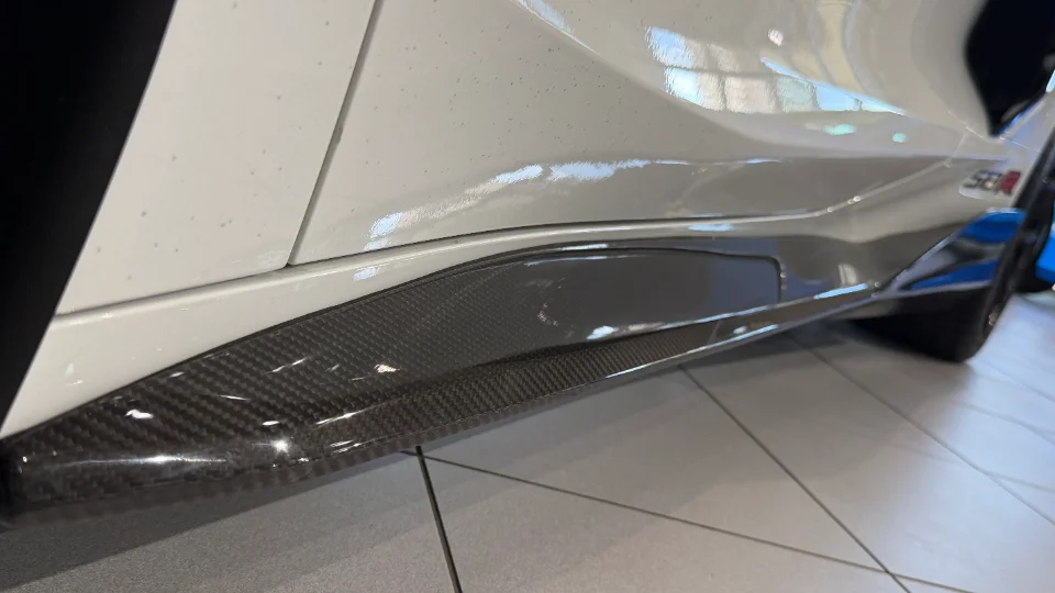 A close-up of a white car's carbon fiber side skirt, showcasing the aerodynamic detail.