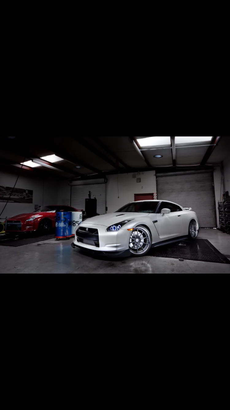 Two Nissan GT-Rs, one white and one red, parked inside a performance tuning shop with a dyno in the background.