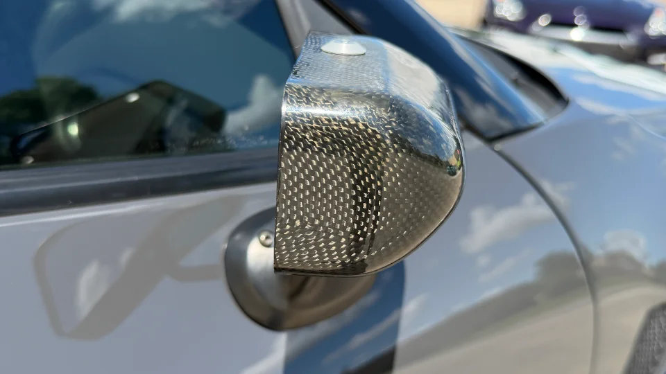 Close-up of a carbon fiber side mirror on a grey performance car, showcasing the weave pattern.