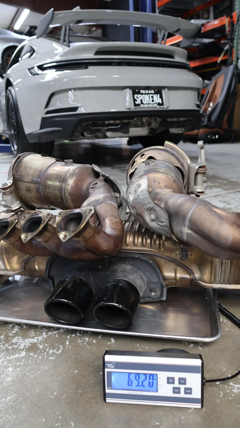 Porsche 911 GT3 on a lift with its OEM exhaust system, including headers and catalytic converters, being weighed on a scale.