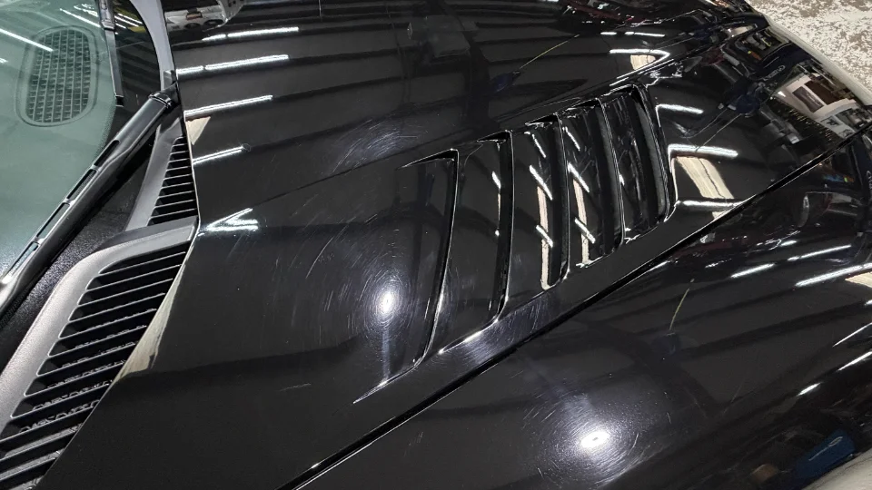 Close-up of a black car hood showing swirl marks and light scratches, indicating a need for paint correction.