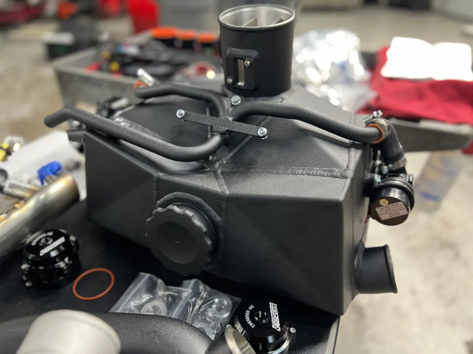 Custom fabricated intercooler or intake plenum with black finish and associated boost components on a workbench in a performance shop.