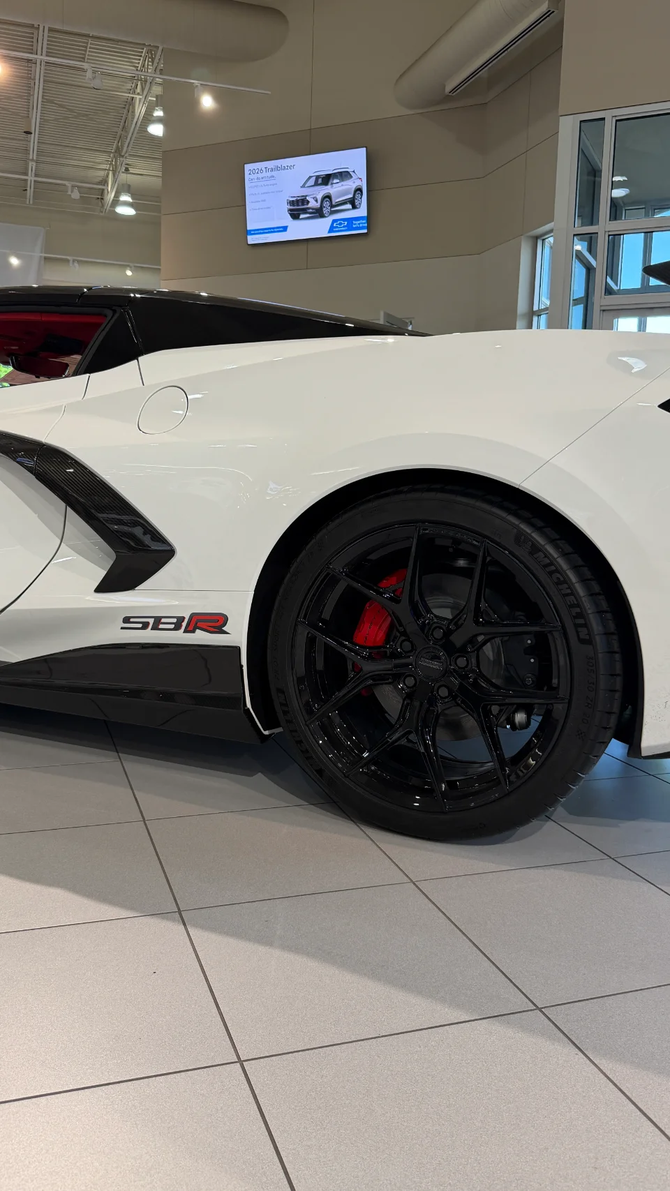 A white Corvette C8 with SBR badging, black aftermarket wheels, and red brake calipers in a showroom.