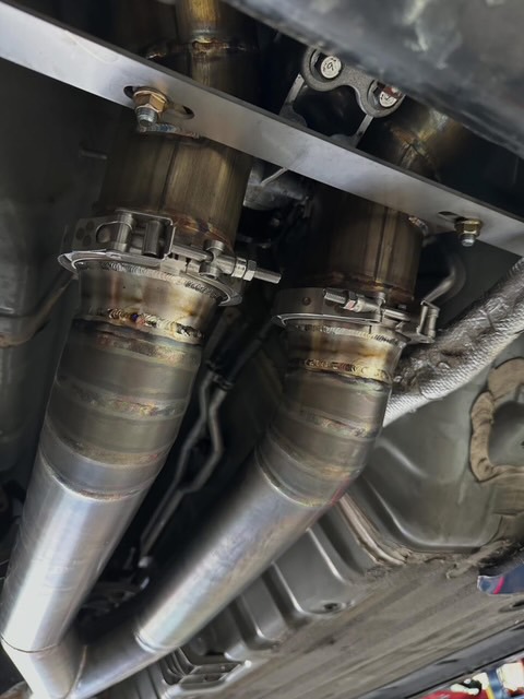 Close-up view of custom stainless steel exhaust downpipes with V-band clamps installed on a performance car's underbody.