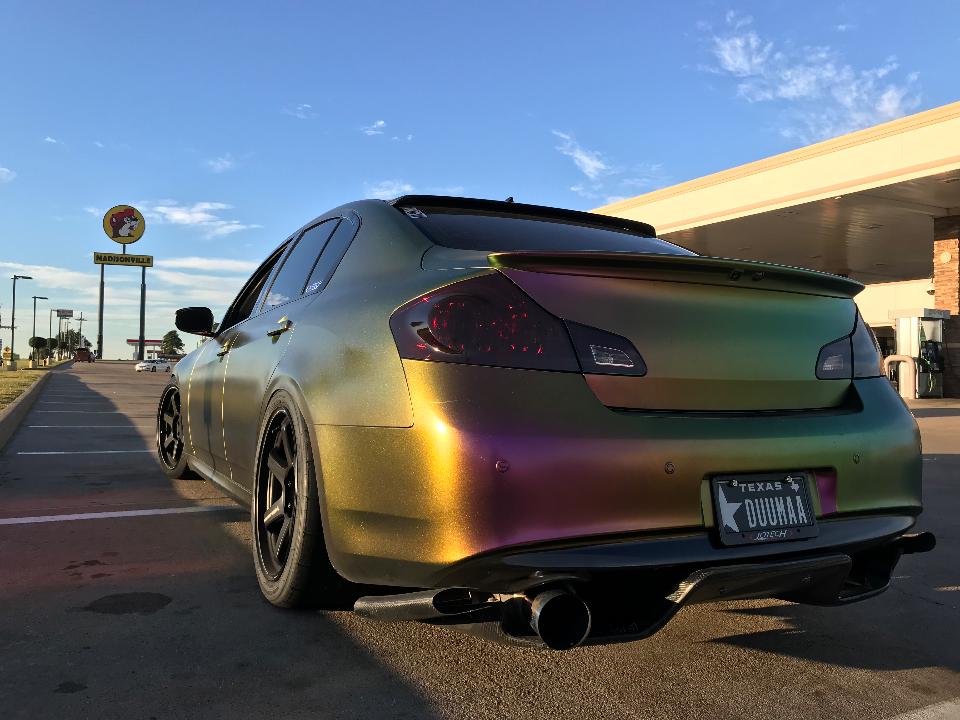 A custom Infiniti G37 sedan with a chameleon wrap, aftermarket exhaust, and rear diffuser parked at a gas station.