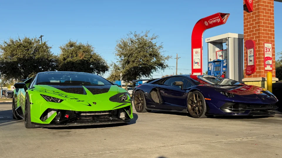 Two Lamborghini supercars, a green Huracan and a blue Aventador, parked at a gas station on a sunny day.