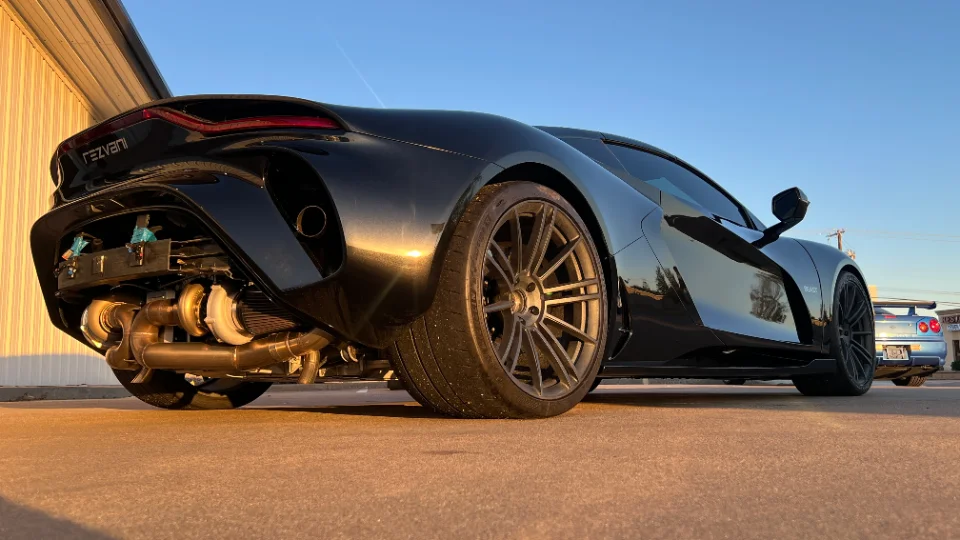 A black Rezvani supercar with exposed twin turbochargers and custom exhaust system, parked outdoors with a blue Nissan GT-R.