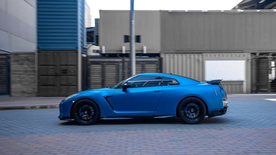 A blue Nissan GT-R R35 driving on a paved street with industrial buildings in the background.